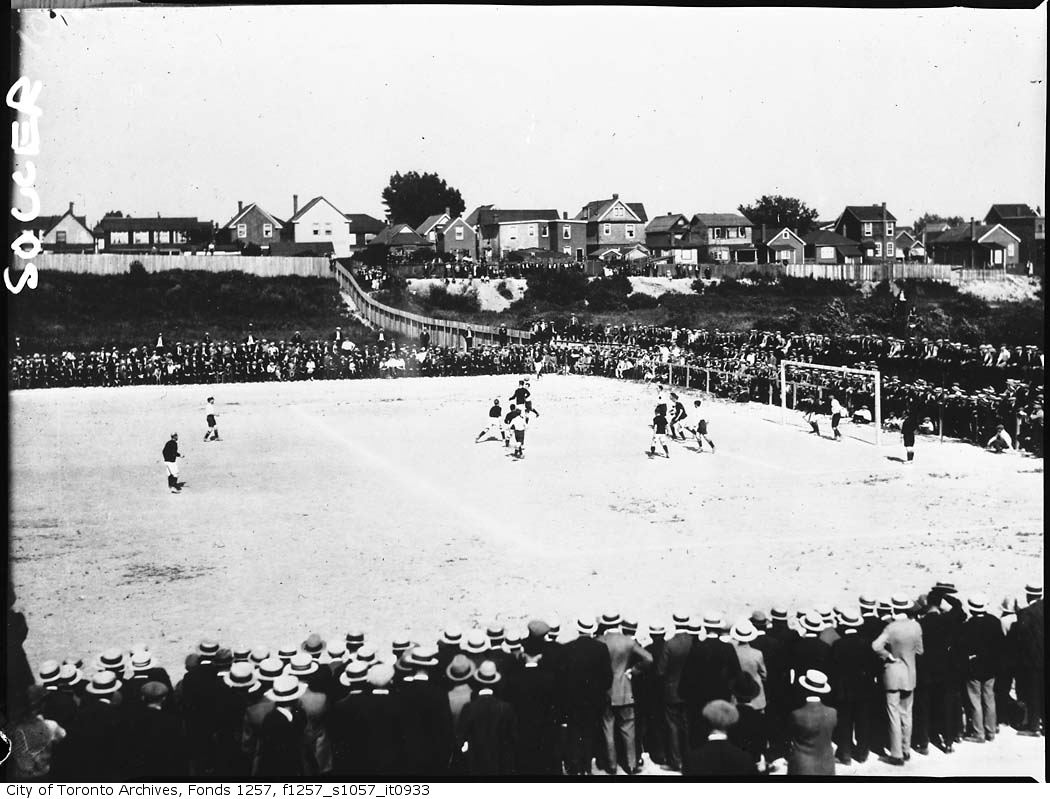 Black and white image of people playing soccer at Ulster Stadium with a row of houses behind the stadium, and fans all around