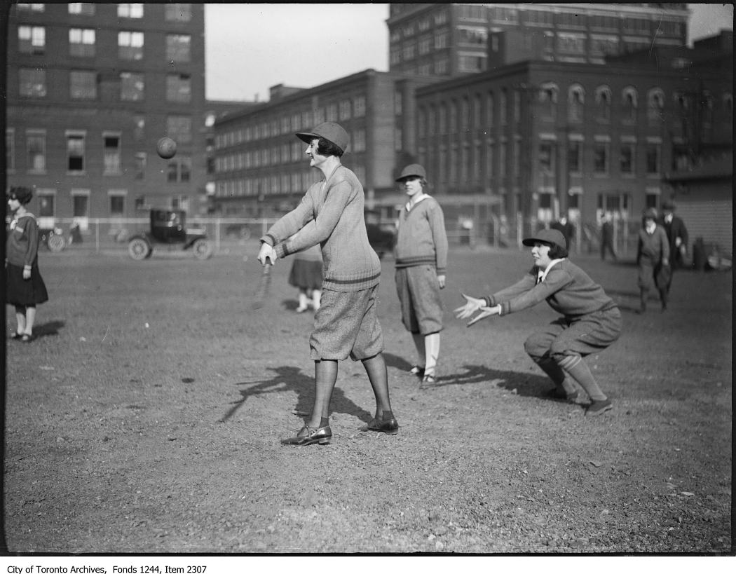 Woman holding a bat as a ball comes towards her, with other women in the background playing softball. All women are wearing ball caps and, sweaters, and bloomers